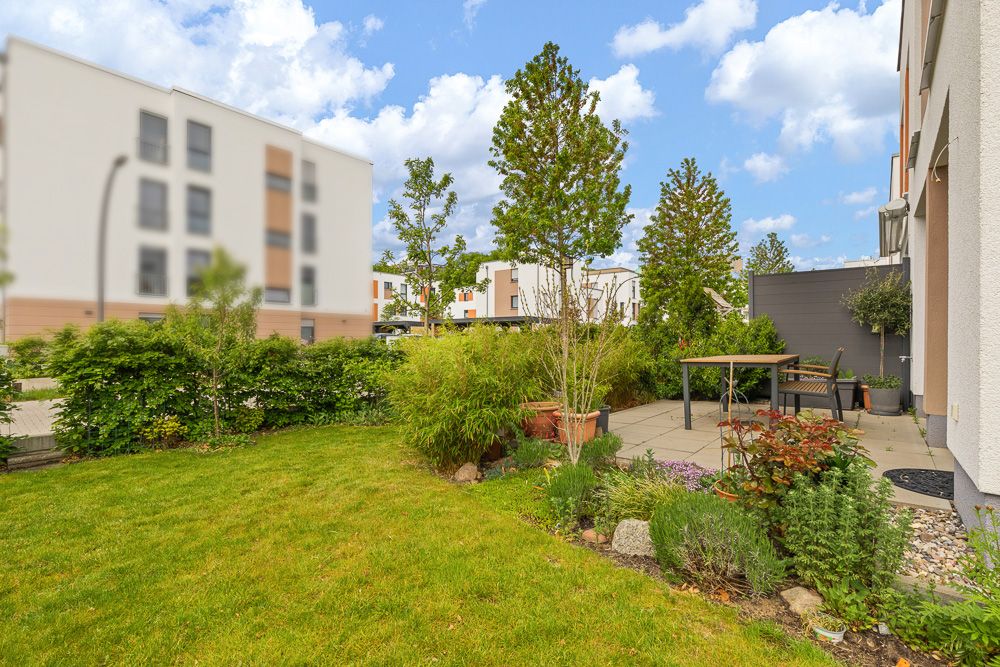 Gepflegter Garten mit Terrasse, Tisch, Stühlen und verschiedenen Pflanzen bei einem Wohnhaus.