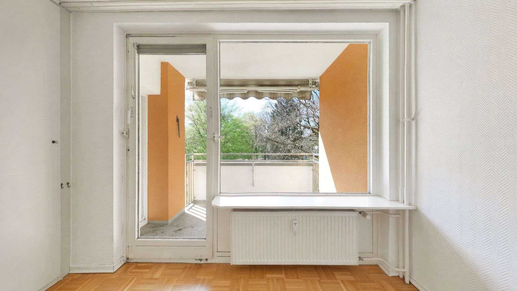 Helles Zimmer mit Parkettboden, großem Fenster und Zugang zu einem Balkon mit Sicht ins Grüne.