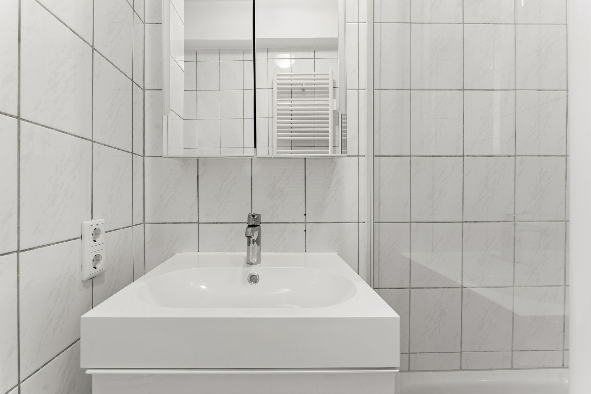 Modernes weiß gefliestes Badezimmer mit Waschbecken, Spiegel und Wasserhahn.