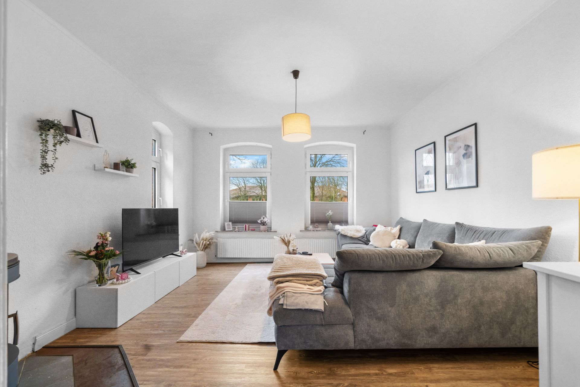 Helles Wohnzimmer mit grauem Sofa, Fernseher, Holzfußboden und drei großen Fenstern.
