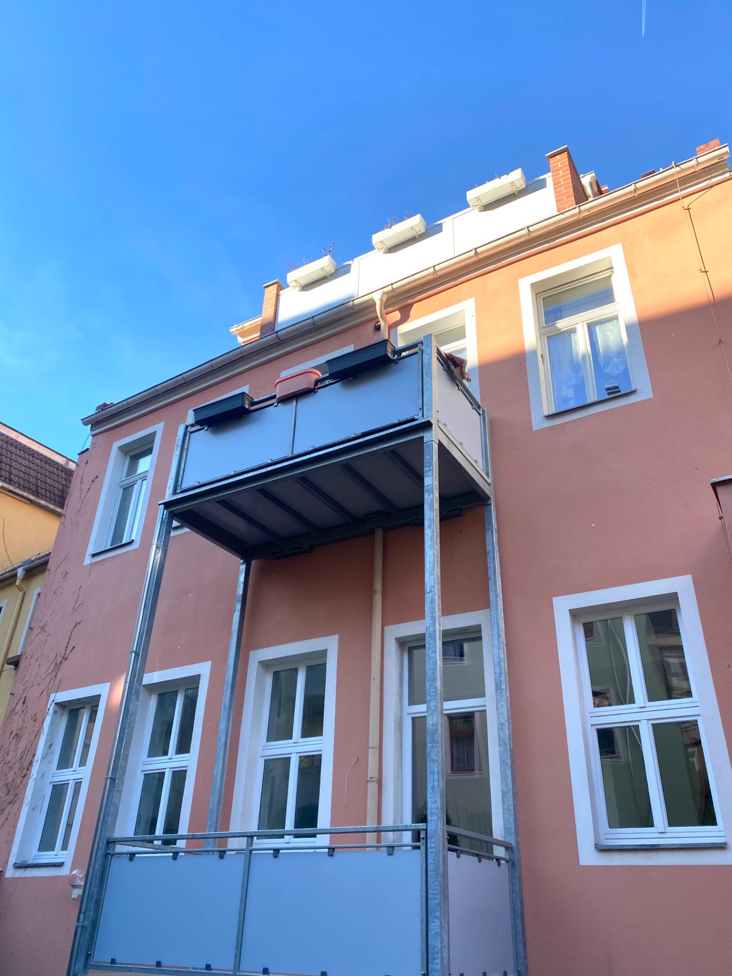 Mehrstöckiges Wohnhaus mit rosa Fassade, weißen Fenstern und zwei Balkonen.