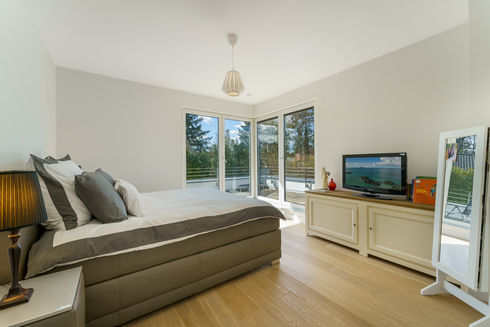 Helles Schlafzimmer mit Doppelbett, großen Fenstern, Kommode mit Fernseher und Stehspiegel.