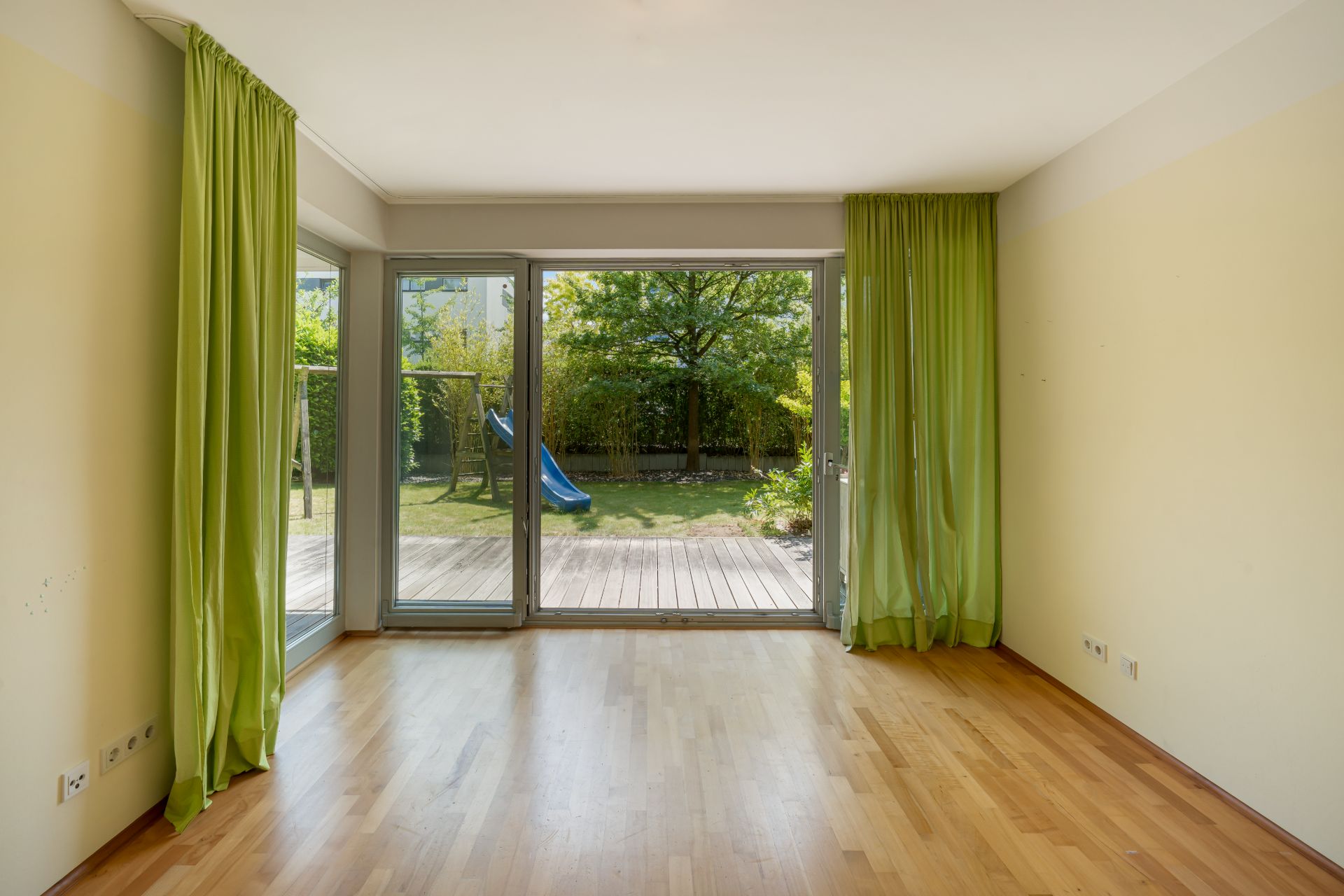 Leeres Zimmer mit Holzboden, großen Fenstern und grünen Vorhängen zum Garten mit Rutsche.