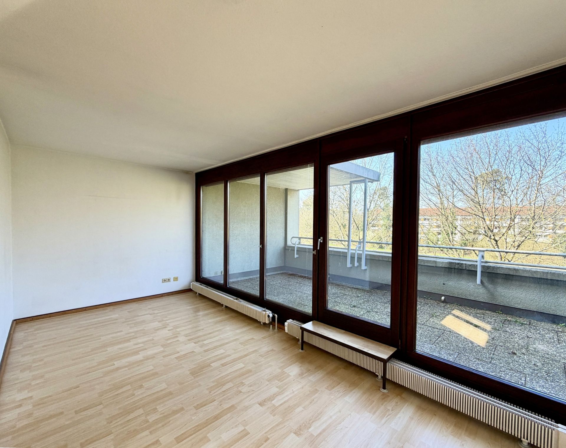 Leerer Raum mit großen Fenstern und Zugang zu einem Balkon, Parkettboden, helle Wände.