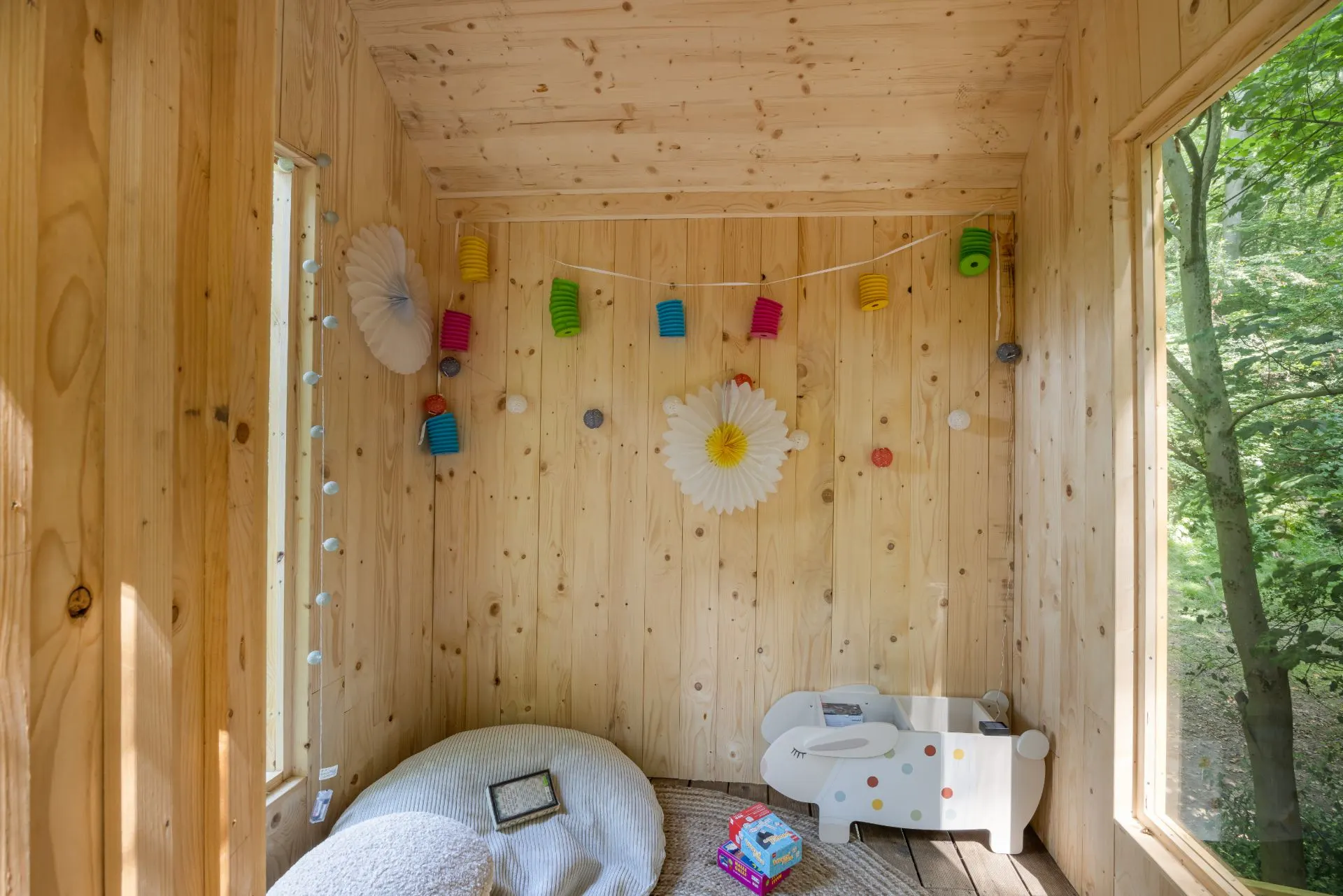 Kleiner Raum mit Holzverkleidung, Spielzeug, Sitzkissen und bunter Papiergirlande an der Wand.