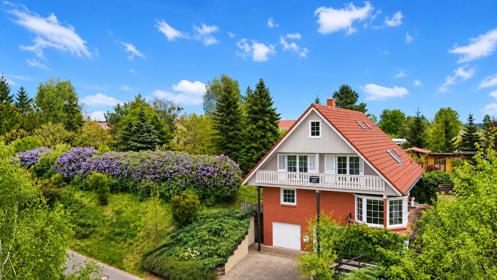 Zweistöckiges rotes Wohnhaus mit Satteldach und großem Balkon, umgeben von Bäumen und Sträuchern.