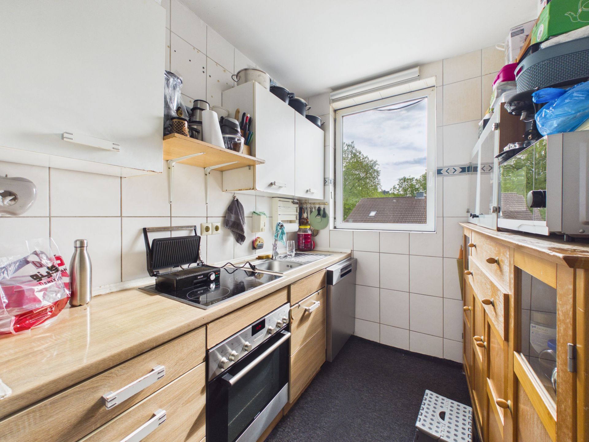 Schmale Küche mit Herd, Spüle, Oberschränken und Fenster mit Blick nach draußen.