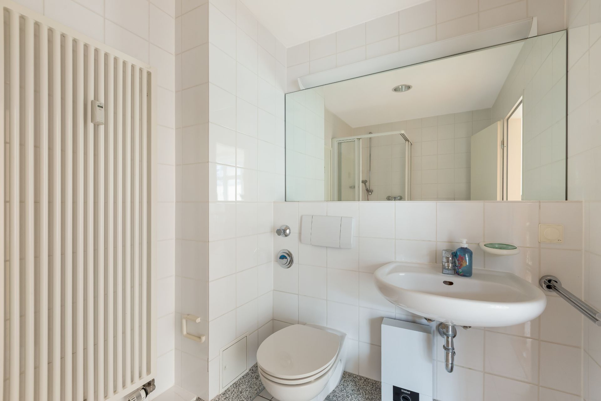 Helles Badezimmer mit WC, Waschbecken, großem Spiegel und Dusche im Hintergrund.