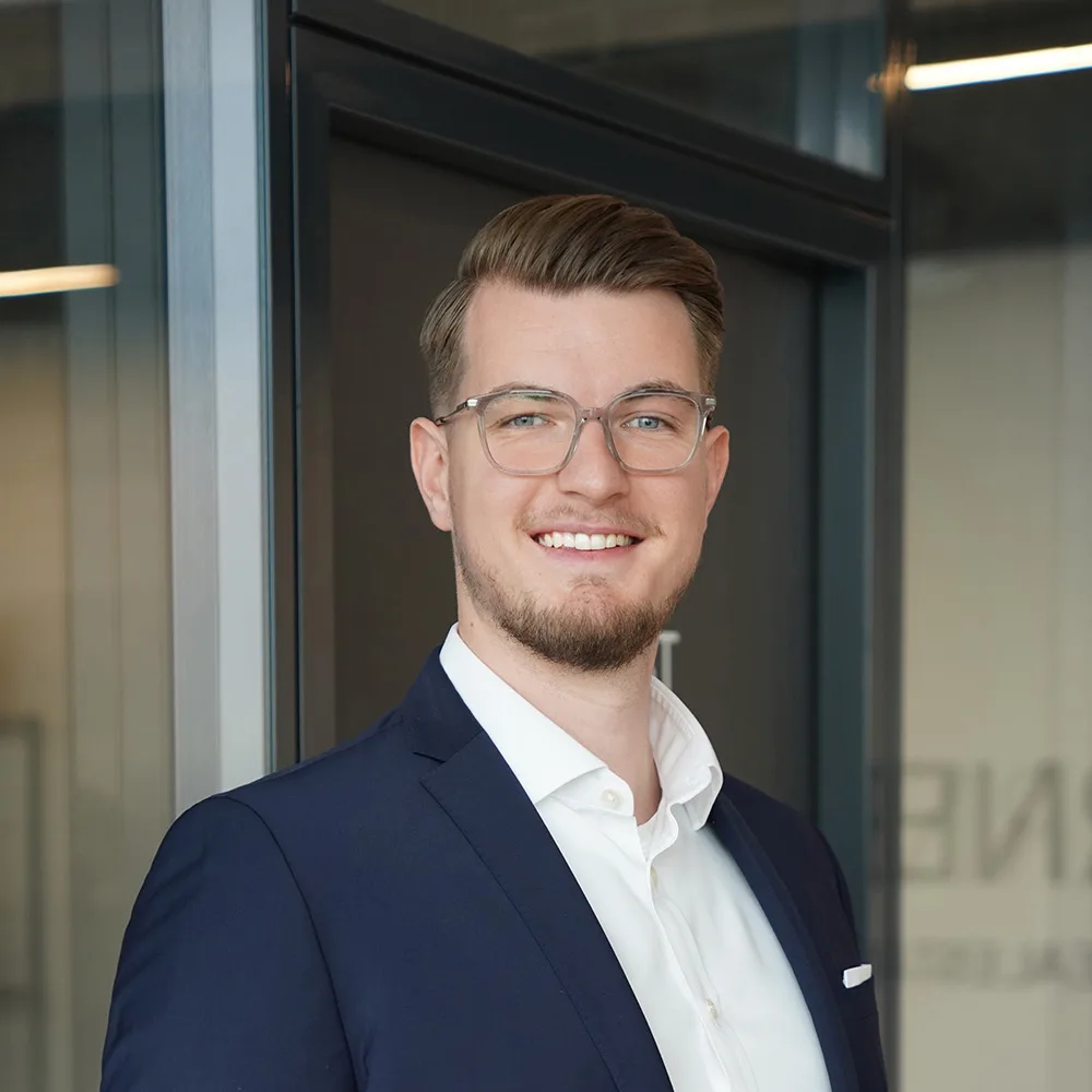Ein Mann mit Brille im Anzug steht vor einer Glastür und lächelt in die Kamera.