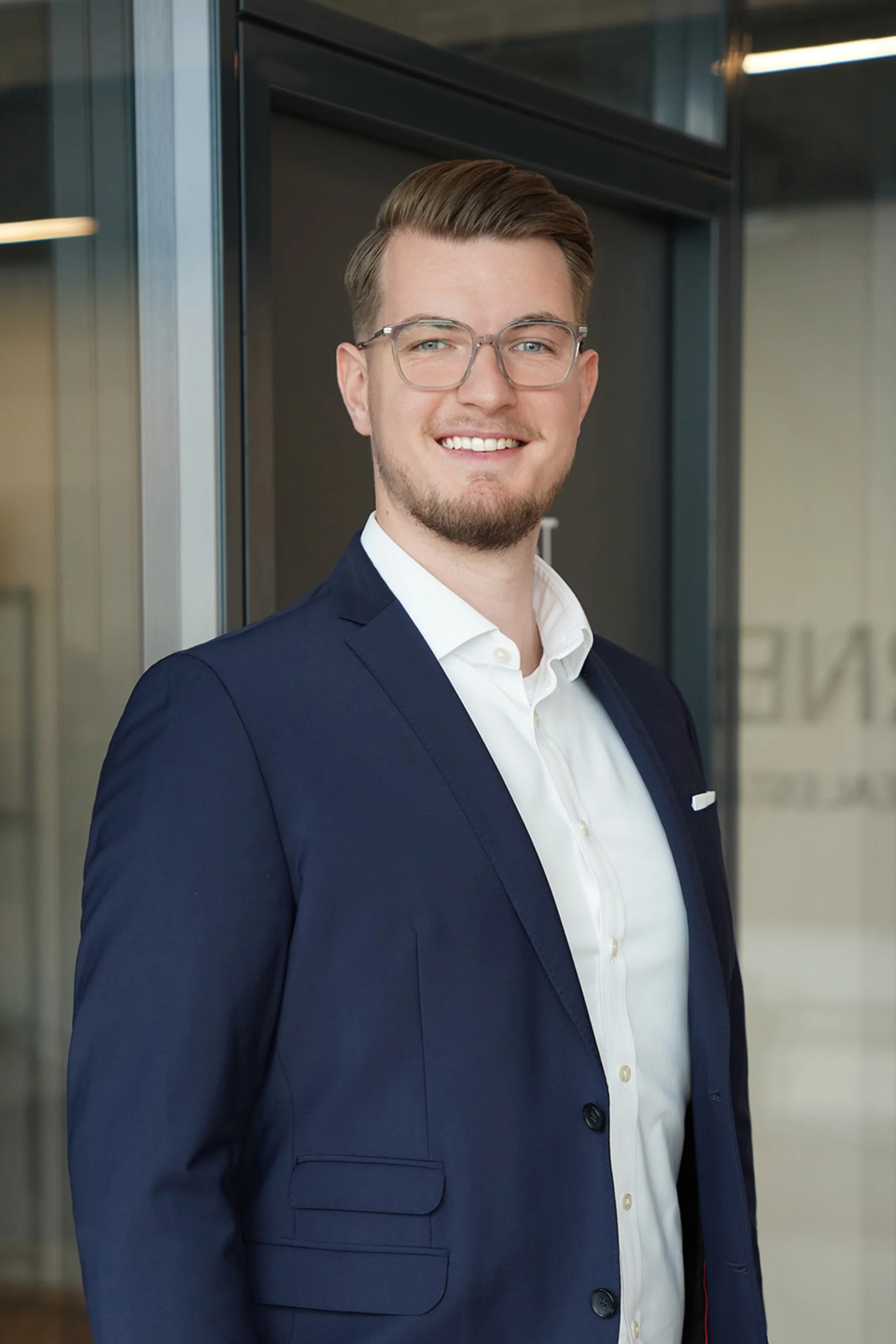 Ein Mann mit Brille im Anzug steht vor einer Glastür und lächelt in die Kamera.
