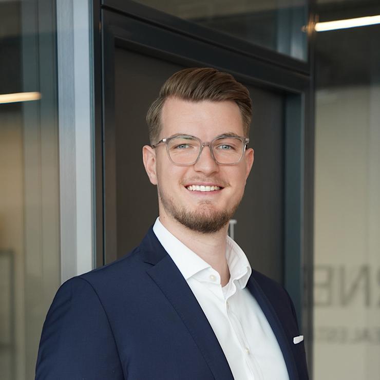 Ein Mann mit Brille im Anzug steht vor einer Glastür und lächelt in die Kamera.