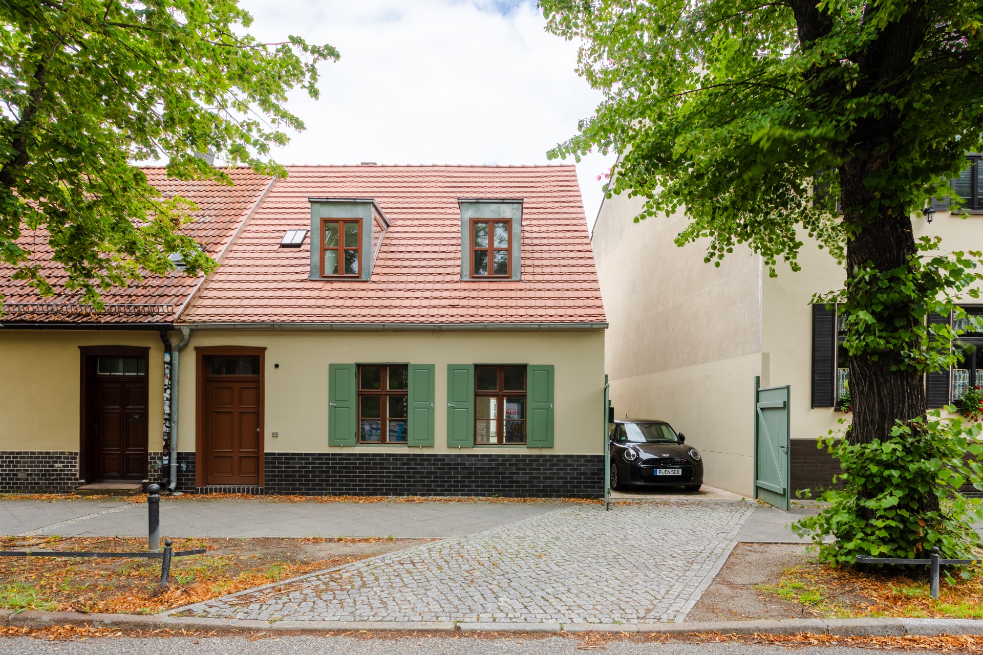 Vorderansicht eines kleinen Hauses mit grünem Fensterladen und Einfahrt mit geparktem Auto.