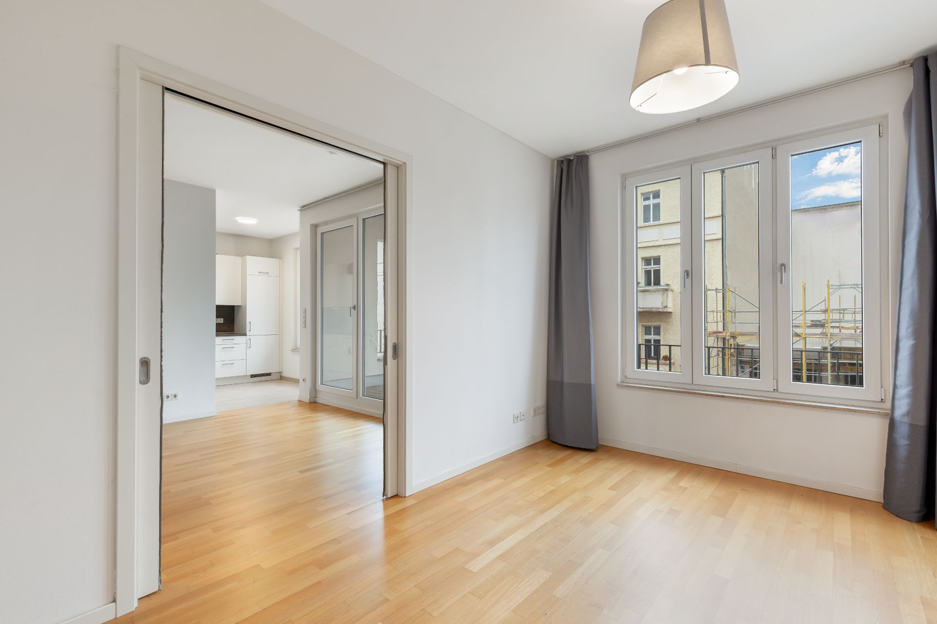 Helles Zimmer mit Holzboden, großem Fenster und Blick in eine offene Küche.