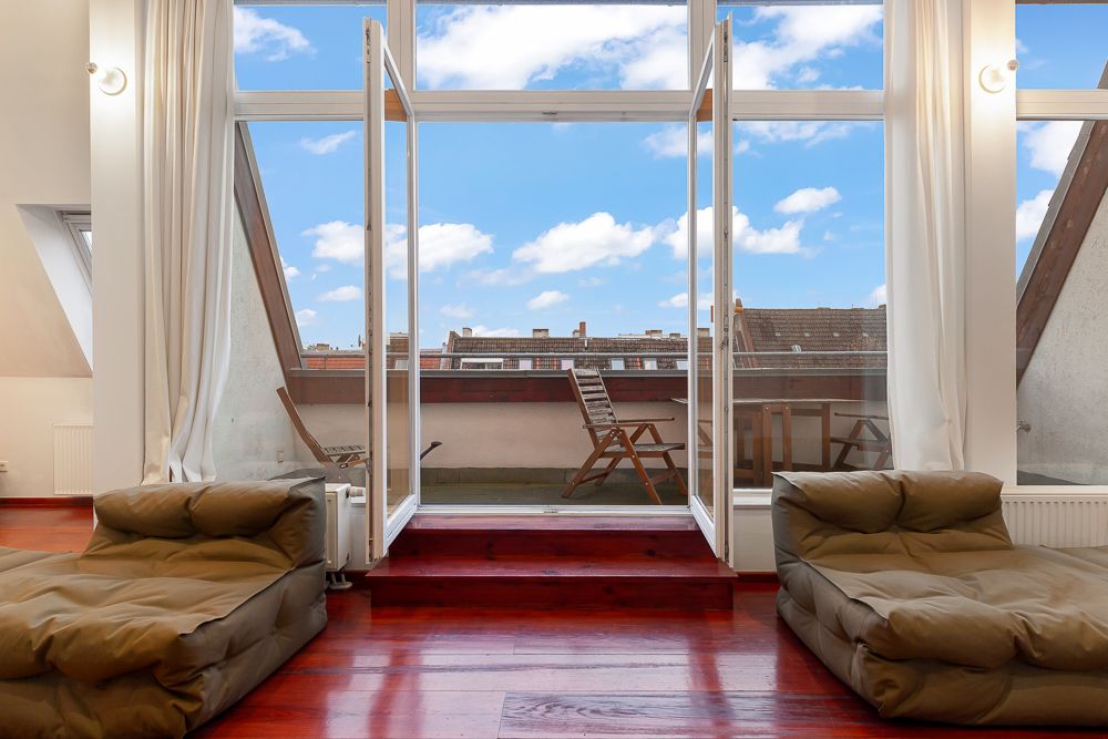 Wohnzimmer mit zwei Sesseln, Holzboden und Zugang zu einem Balkon mit Tisch und Stühlen.