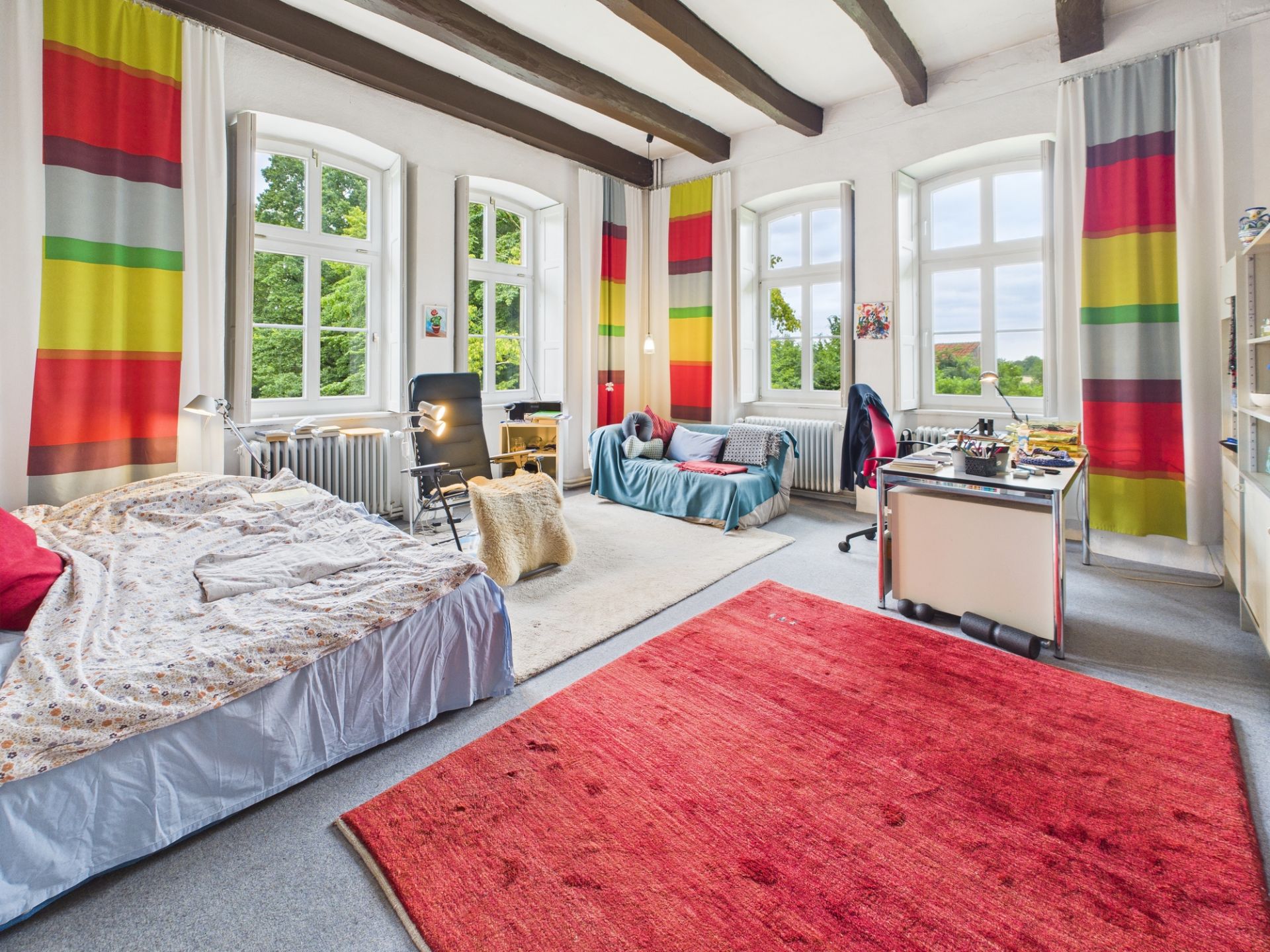 Helles Schlafzimmer mit Bett, Schreibtisch, Sofa und bunten Vorhängen vor großen Fenstern.