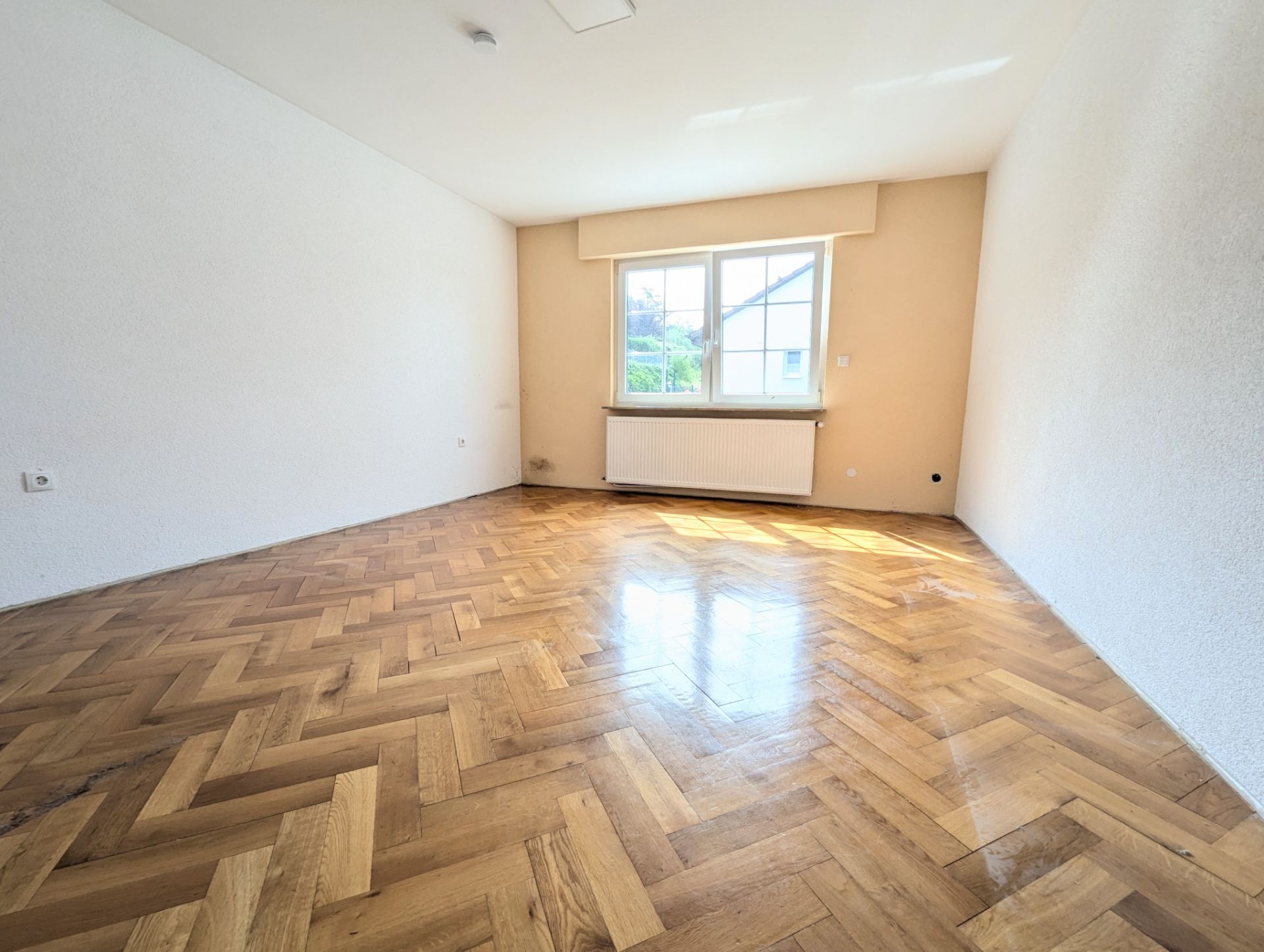 Leerer Raum mit Fischgrätparkettboden und großem Fenster.