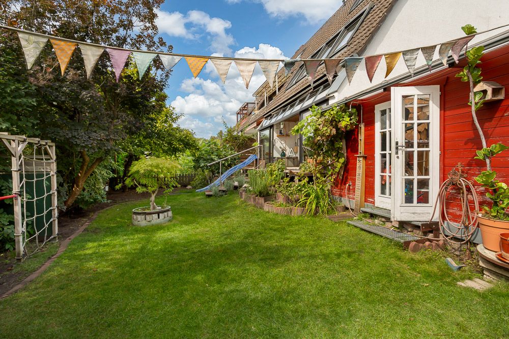 Grüner Garten mit Wimpelkette, roter Hauswand, Glastür und Spielgeräten bei sonnigem Wetter.