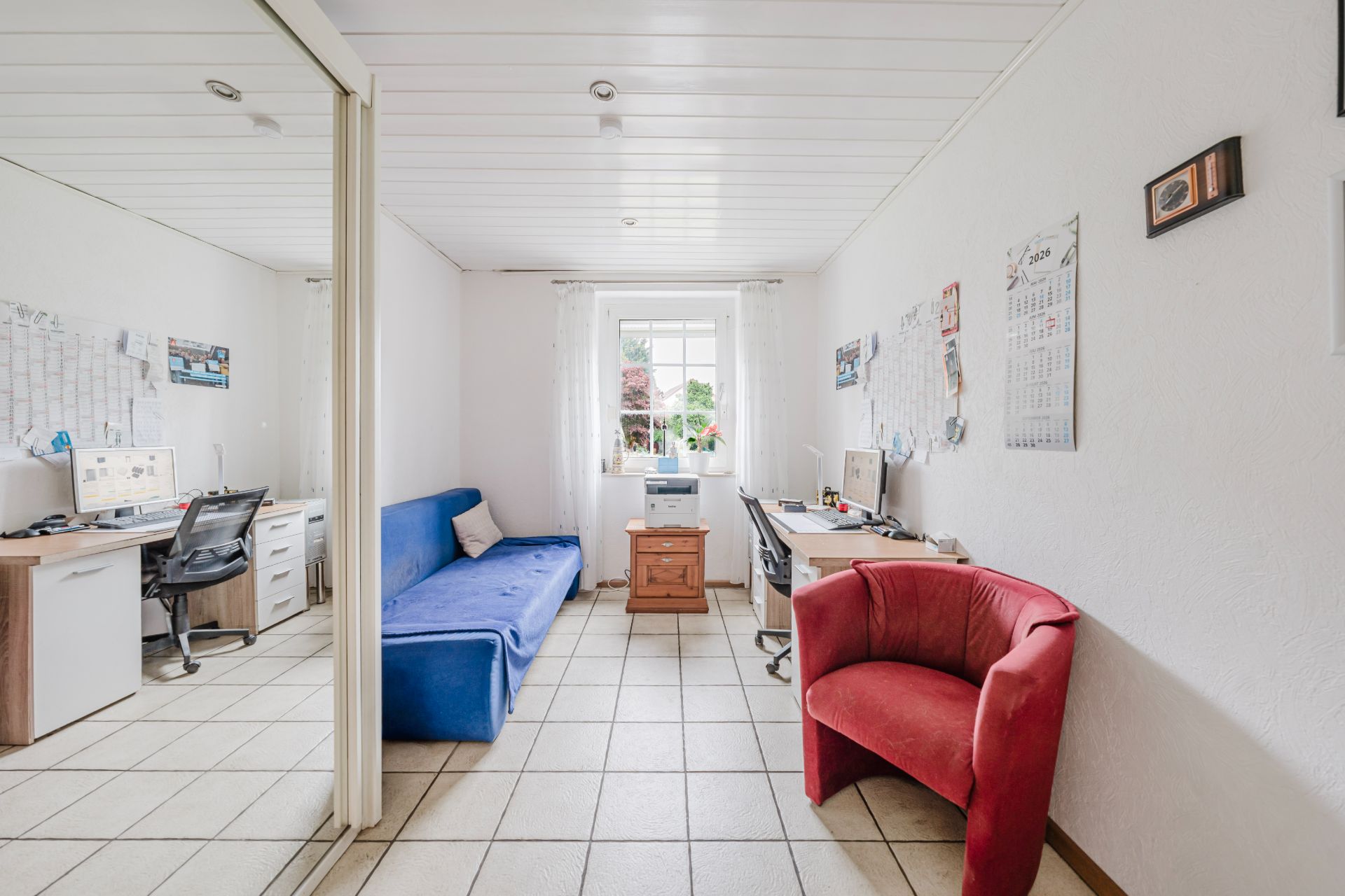 Helles Arbeitszimmer mit Schreibtischen, blauem Sofa, rotem Sessel und Fenster.