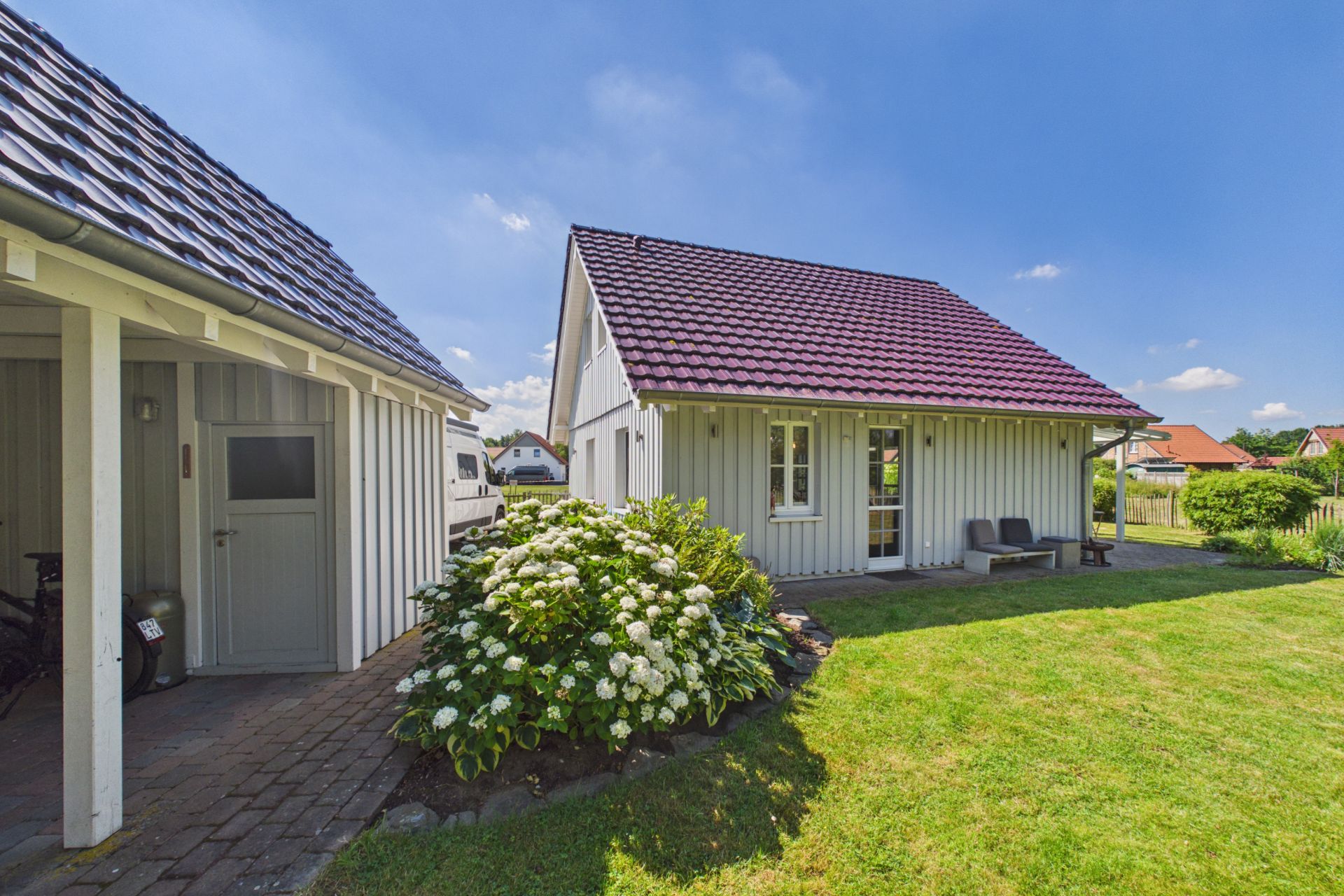 Weißes Haus mit rotem Dach, Garten, Bank und blühenden Sträuchern im Vordergrund.