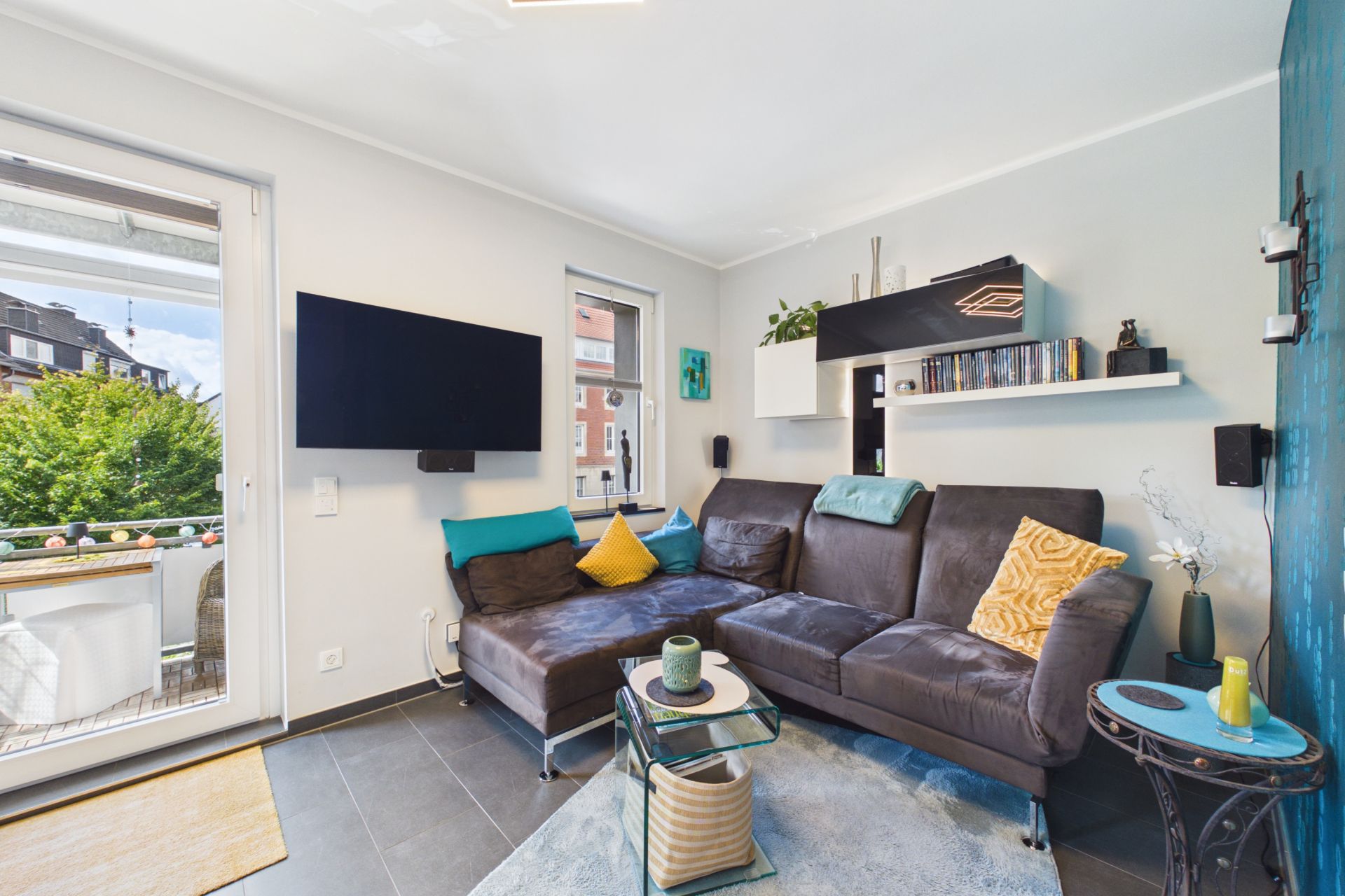 Modernes Wohnzimmer mit Ecksofa, Wandregalen, Fernseher und Zugang zu einem Balkon.