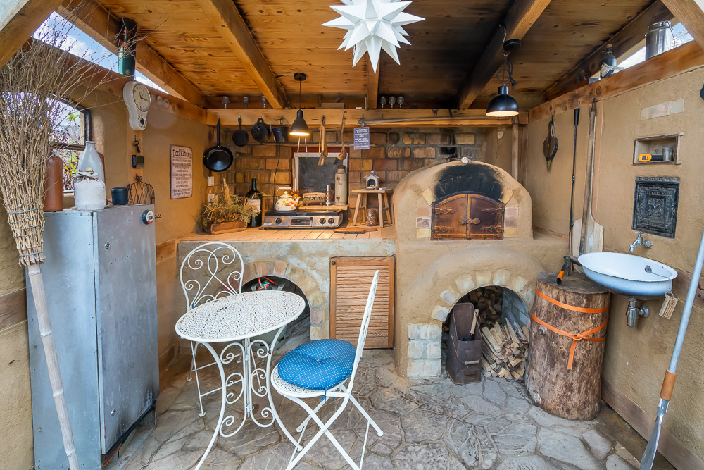 Überdachte Außenküche mit Holzofen, kleinem Tisch und Stühlen auf Steinboden.