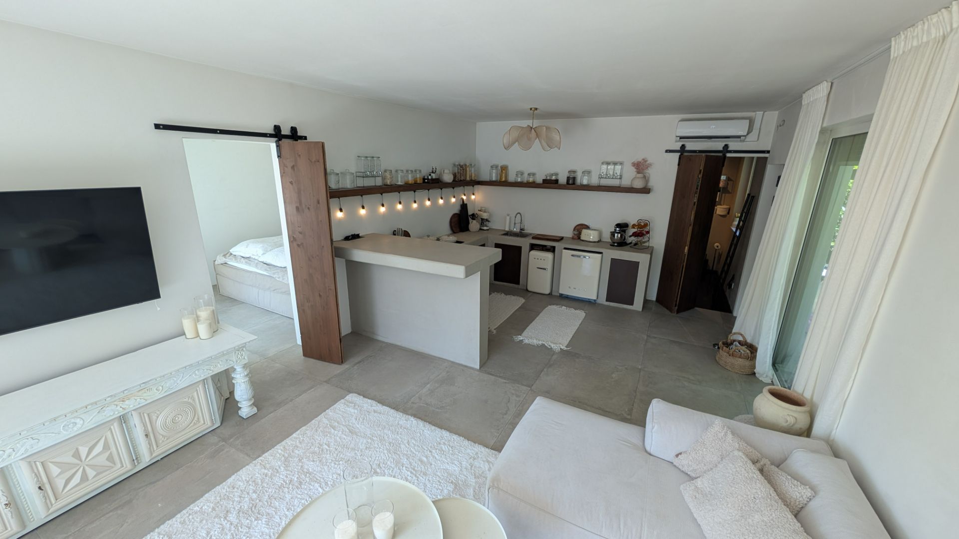 Helles Wohnzimmer mit Sofa, TV, offener Küche und sichtbarem Schlafzimmerbereich.