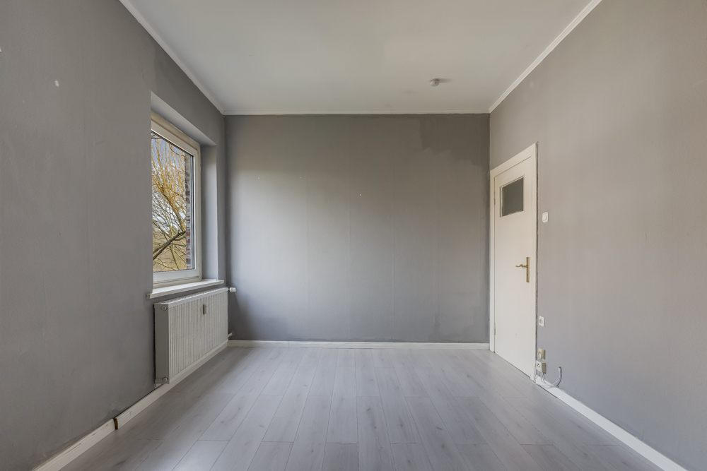 Leerer Raum mit grauen Wänden, Fenster links und weißer Tür rechts.