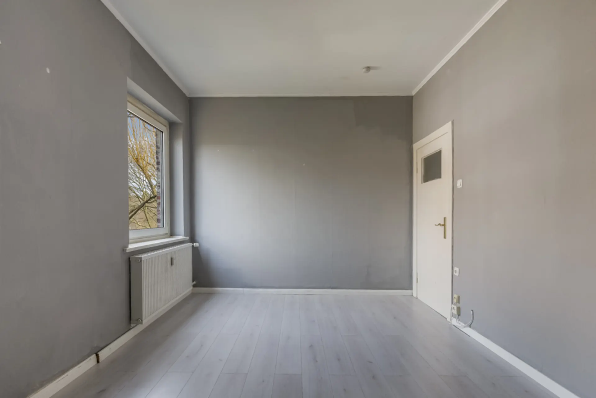 Leerer Raum mit grauen Wänden, Fenster links und weißer Tür rechts.