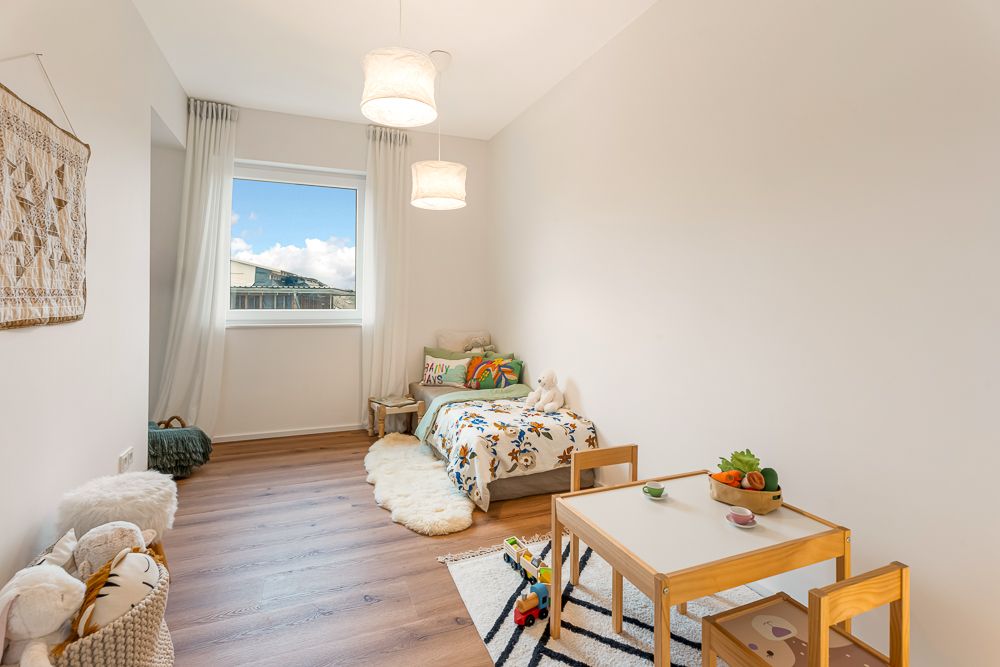Helles Kinderzimmer mit Einzelbett, kleinem Tisch, zwei Stühlen und Spielzeug.