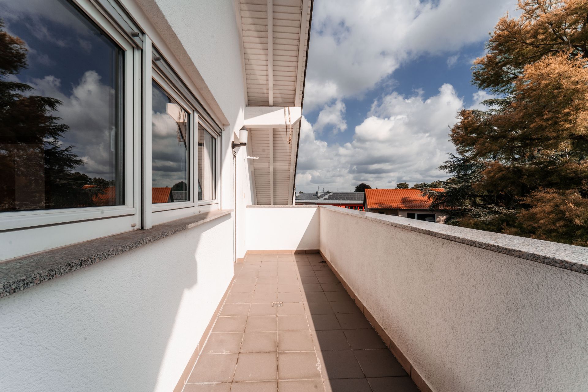 Langer Balkon mit Fliesenboden, weißen Wänden und Blick auf Bäume und Dächer.