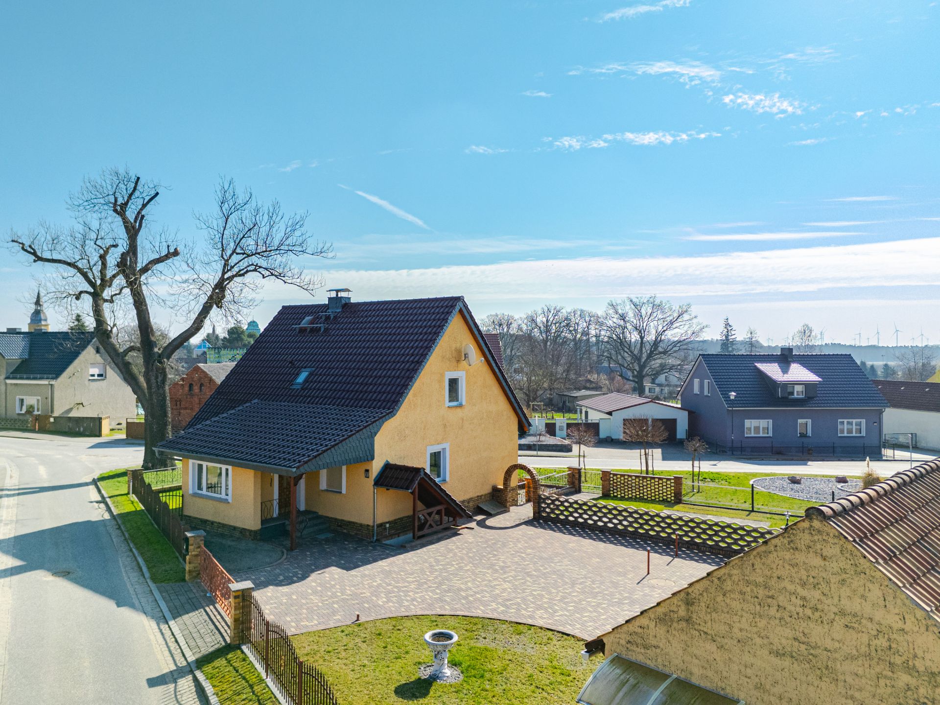 Gelbes Einfamilienhaus mit dunklem Dach, gepflasterter Hof, Bäumen und Nachbarhäusern an einer Straße.