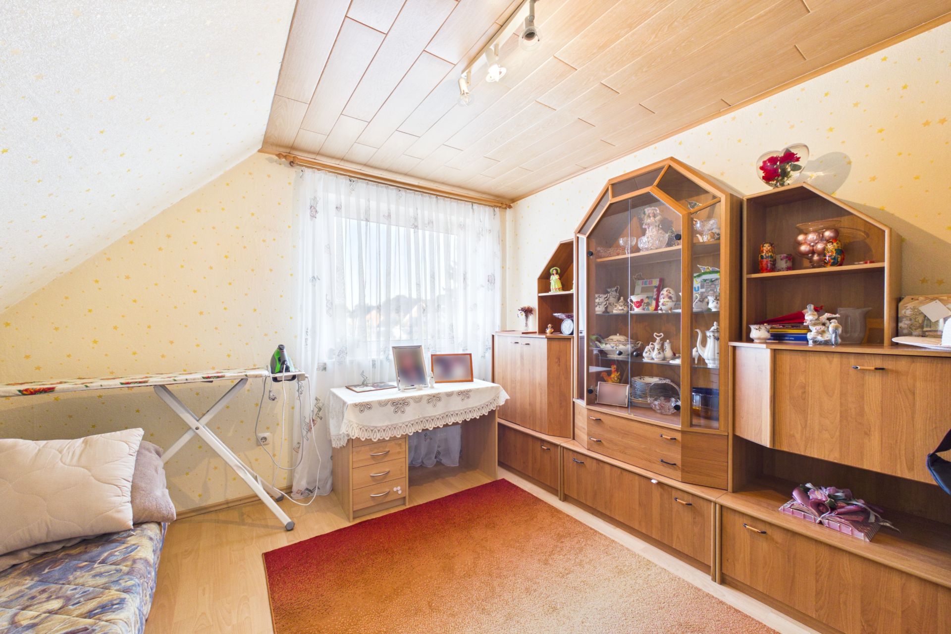 Helles Zimmer mit Schrankwand, Sofa, Bügelbrett und großem Fenster mit Gardine.