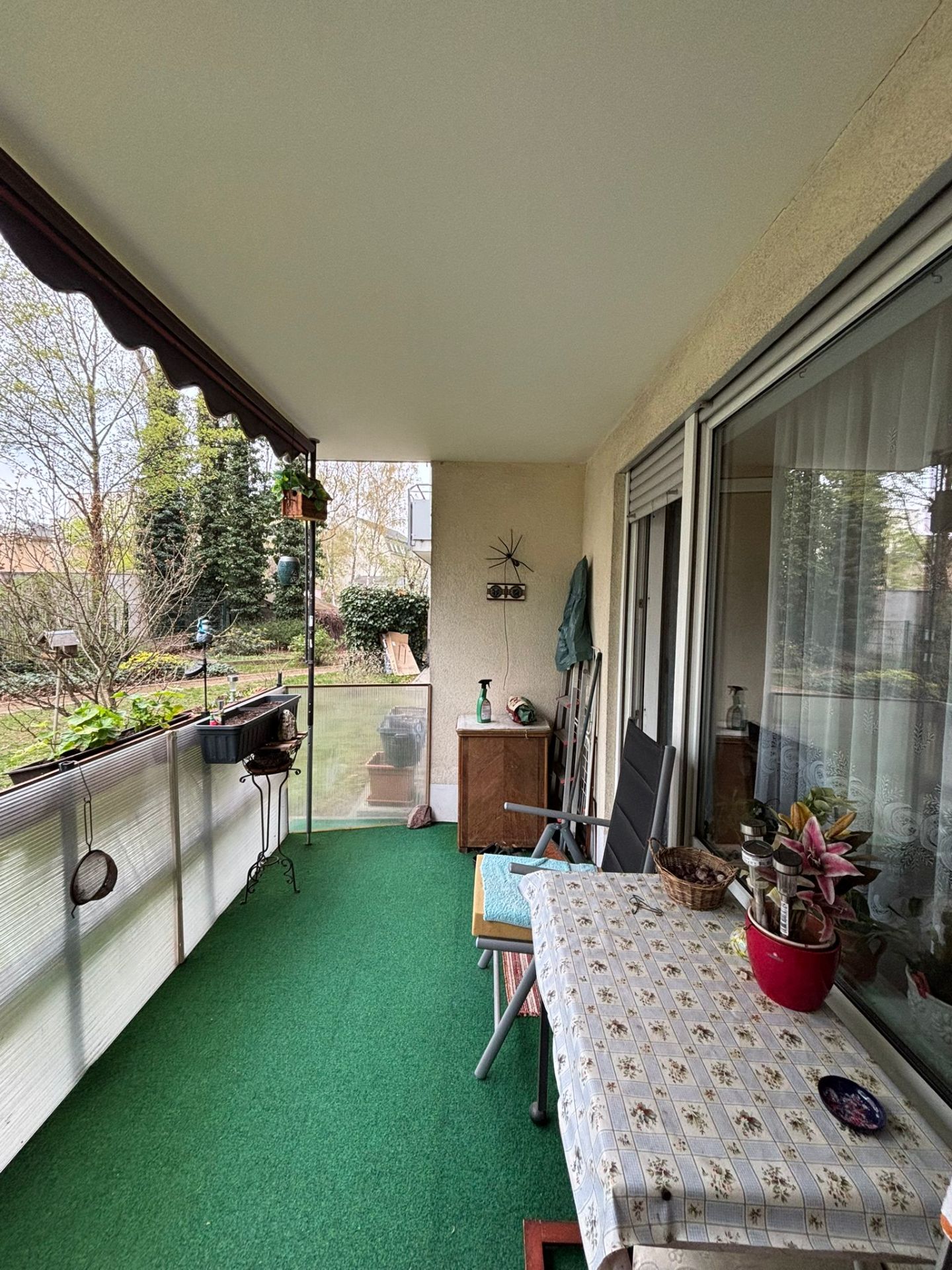 Überdachter Balkon mit grünem Teppich, Tisch, Stühlen und Pflanzen, Blick auf einen Garten.