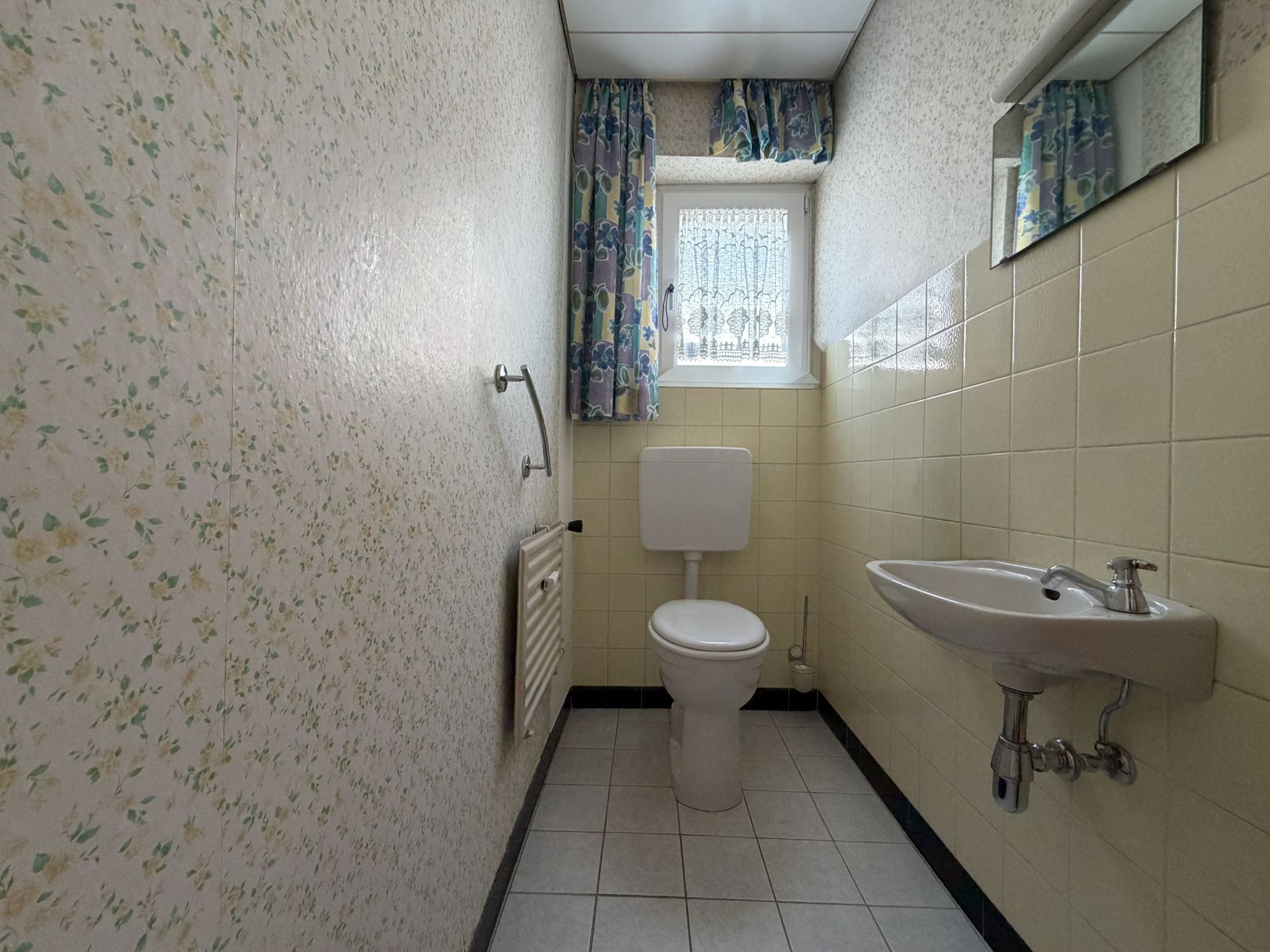 Kleines Bad mit Toilette, Waschbecken, Fenster mit Vorhang und Spiegel.
