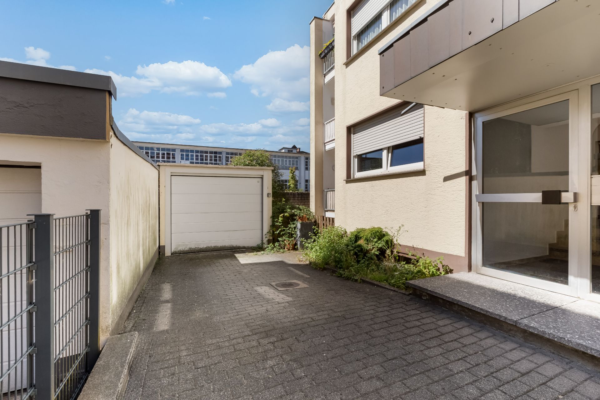 Einfahrt mit Garage und Eingangstür neben einem Wohnhaus, einige Pflanzen am Rand.