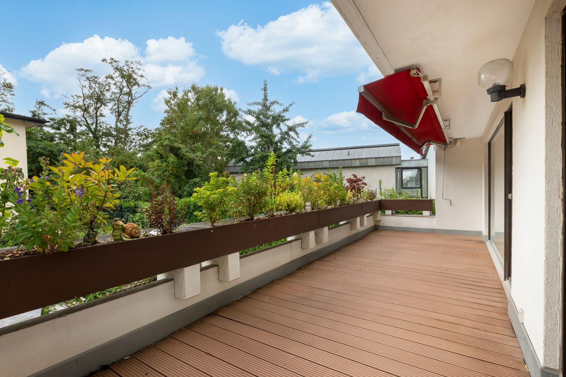 Großer Balkon mit Holzboden, Blumenkästen und roter Markise, Blick ins Grüne.
