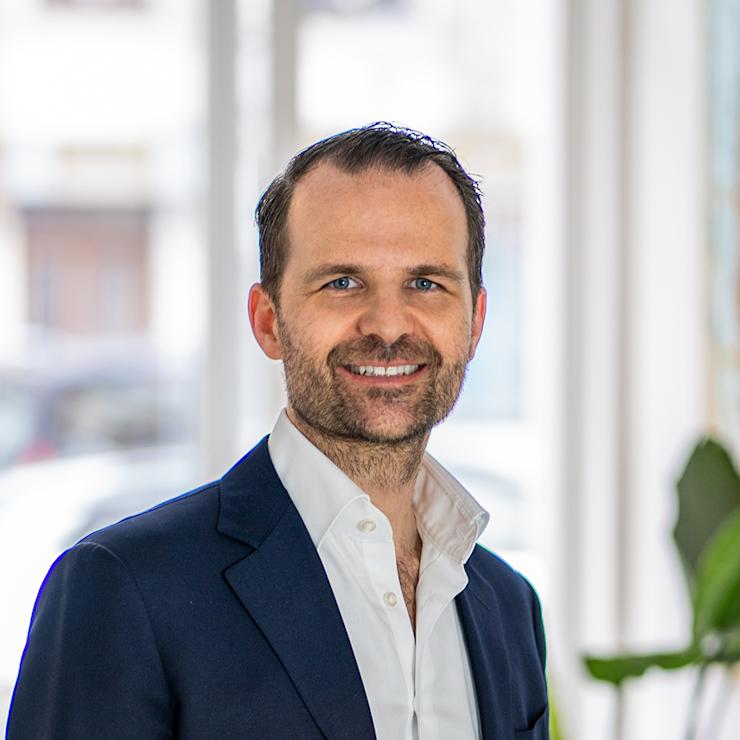 Ein Mann im Anzug steht in einem hellen Raum mit Fenstern und Pflanzen im Hintergrund.