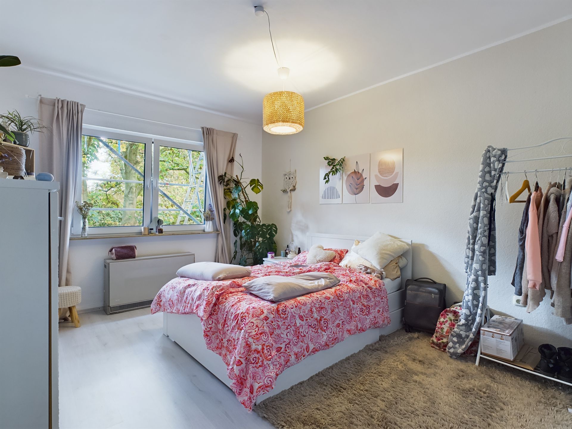 Helles Schlafzimmer mit Doppelbett, Kleiderständer, Teppich und großem Fenster mit Blick ins Grüne.