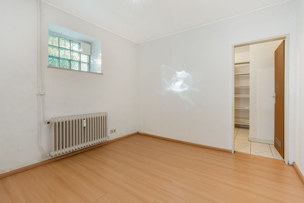 Leeres Zimmer mit Laminatboden, Heizkörper, kleinem Fenster und Tür zu einem Raum mit Regalen.