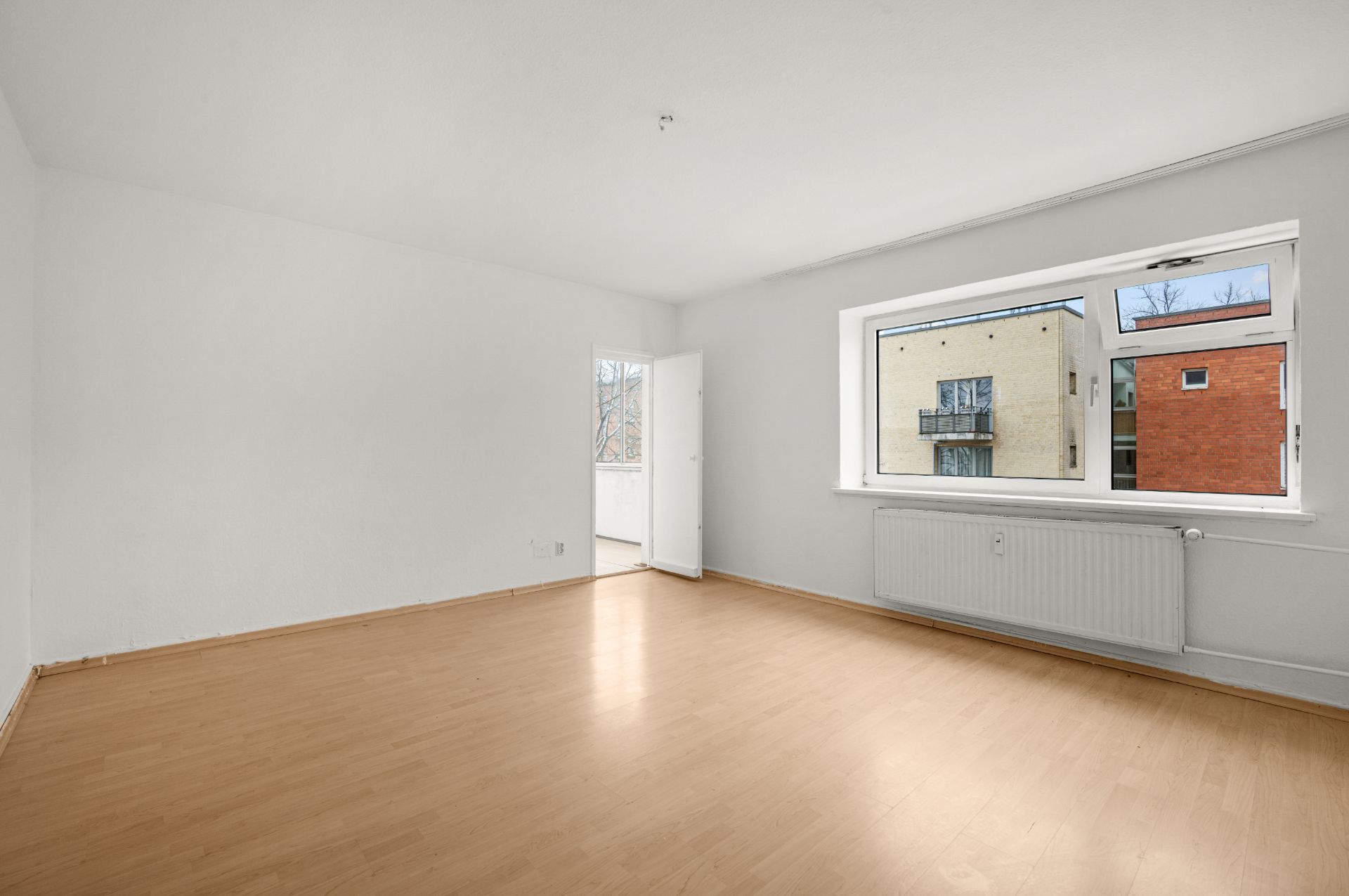 Heller, leerer Raum mit Holzfußboden, weiß gestrichenen Wänden und großem Fenster.