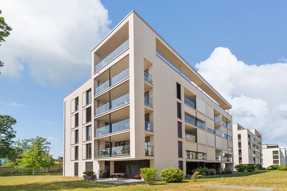 Modernes Mehrfamilienhaus mit Balkonen, umgeben von Grünflächen und Bäumen.