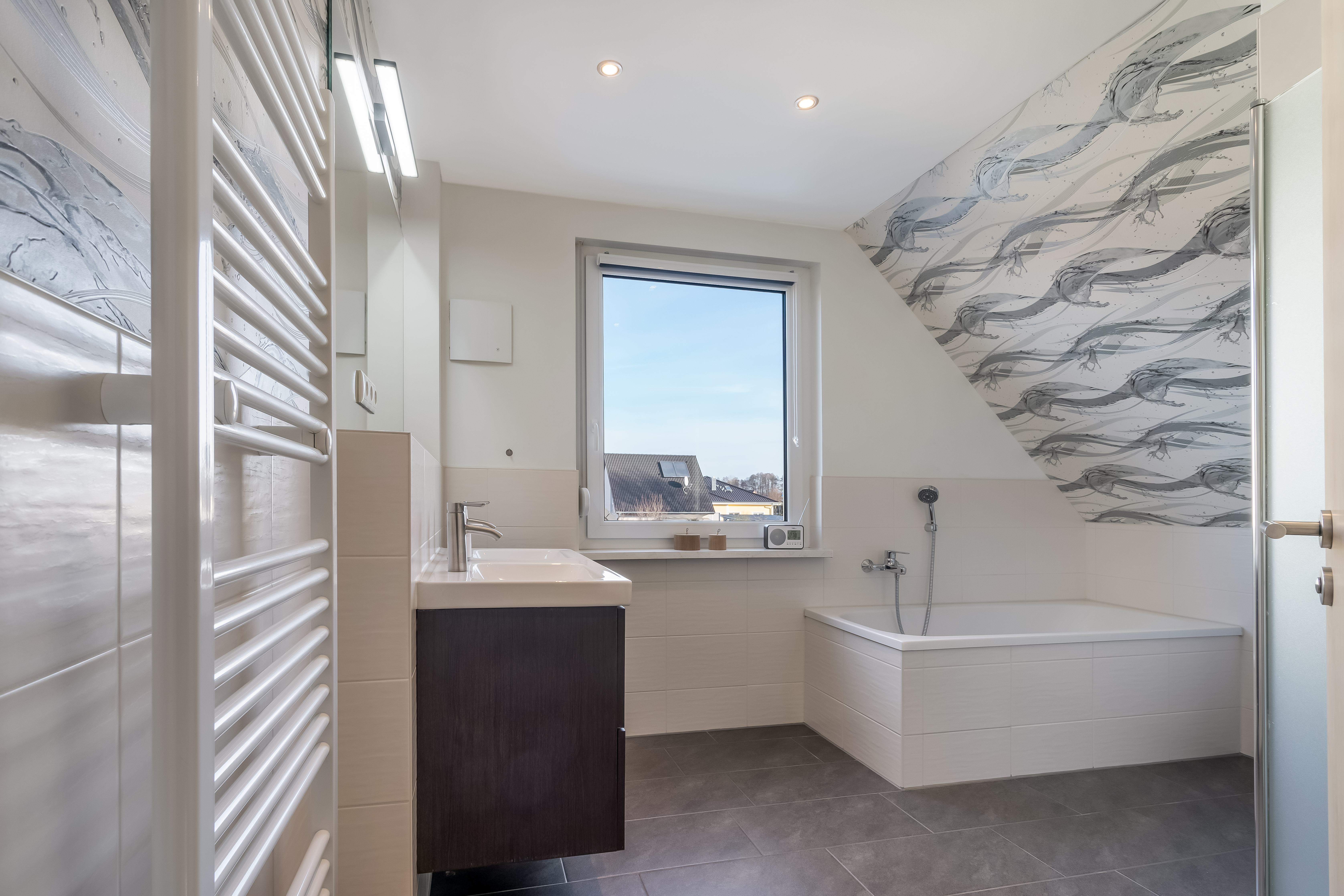 Helles Badezimmer mit Badewanne, großem Fenster, Waschbecken, Handtuchheizkörper und gemusterter Wand.