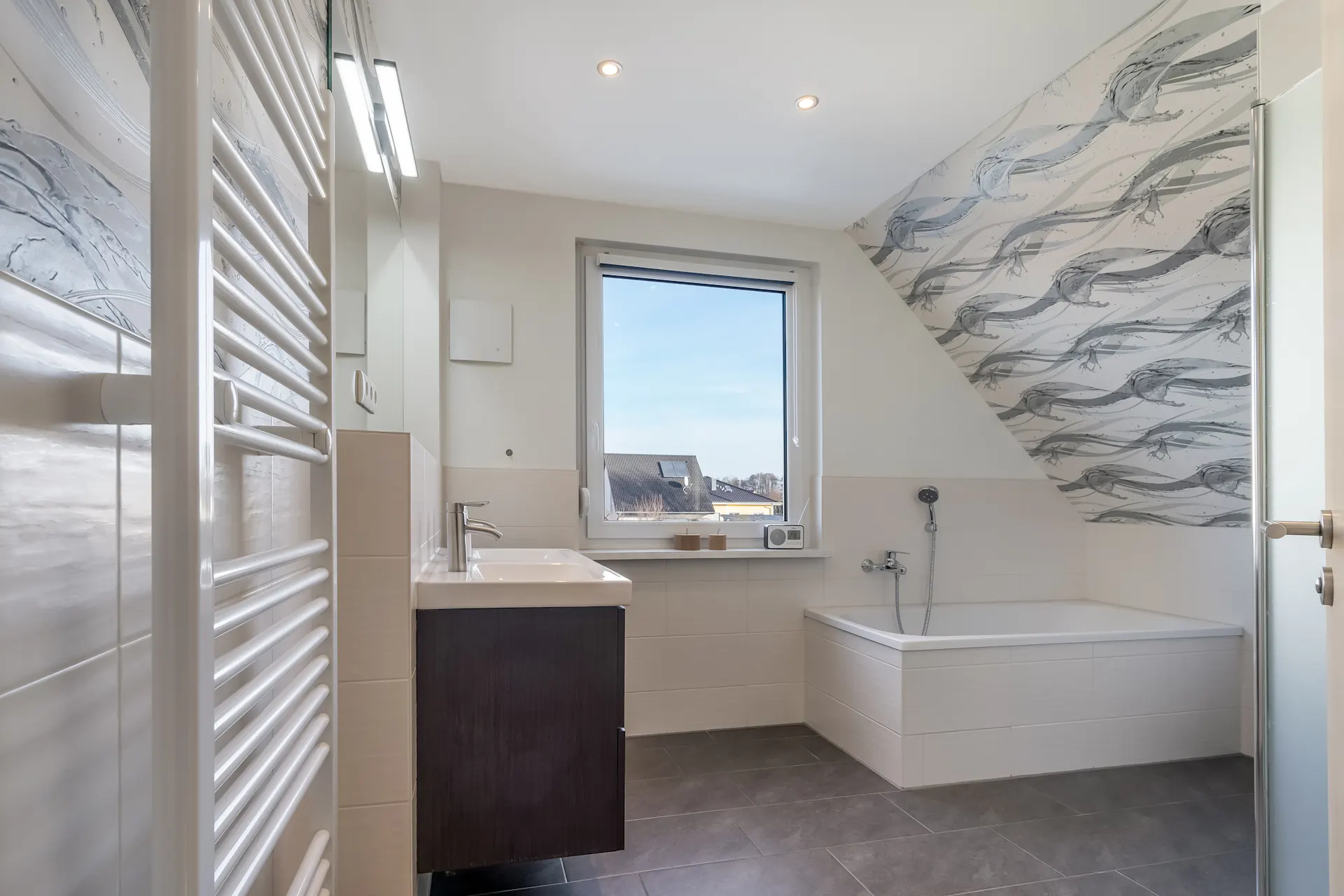 Helles Badezimmer mit Badewanne, großem Fenster, Waschbecken, Handtuchheizkörper und gemusterter Wand.