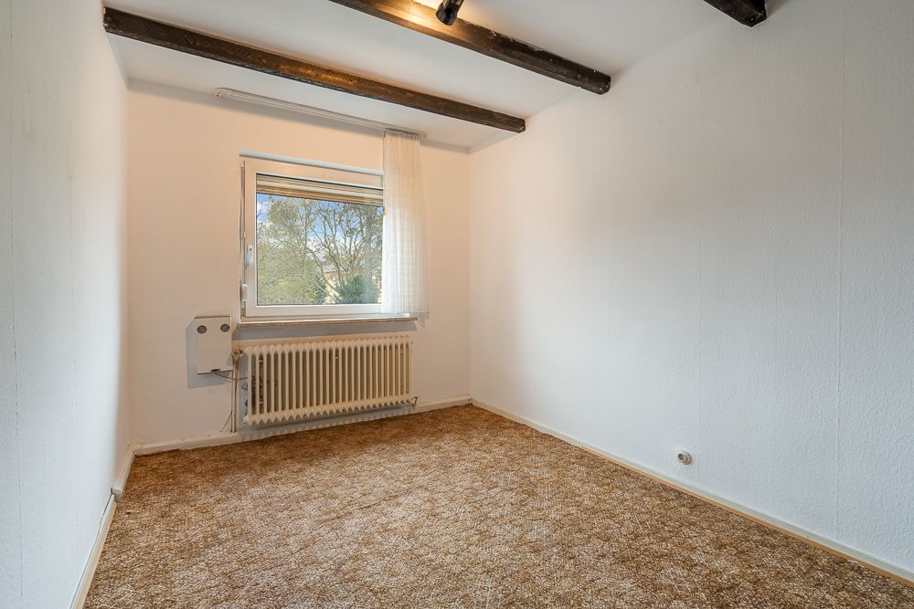 Leeres Zimmer mit braunem Teppich, weißem Heizkörper, Fenster und Holzbalken an der Decke.
