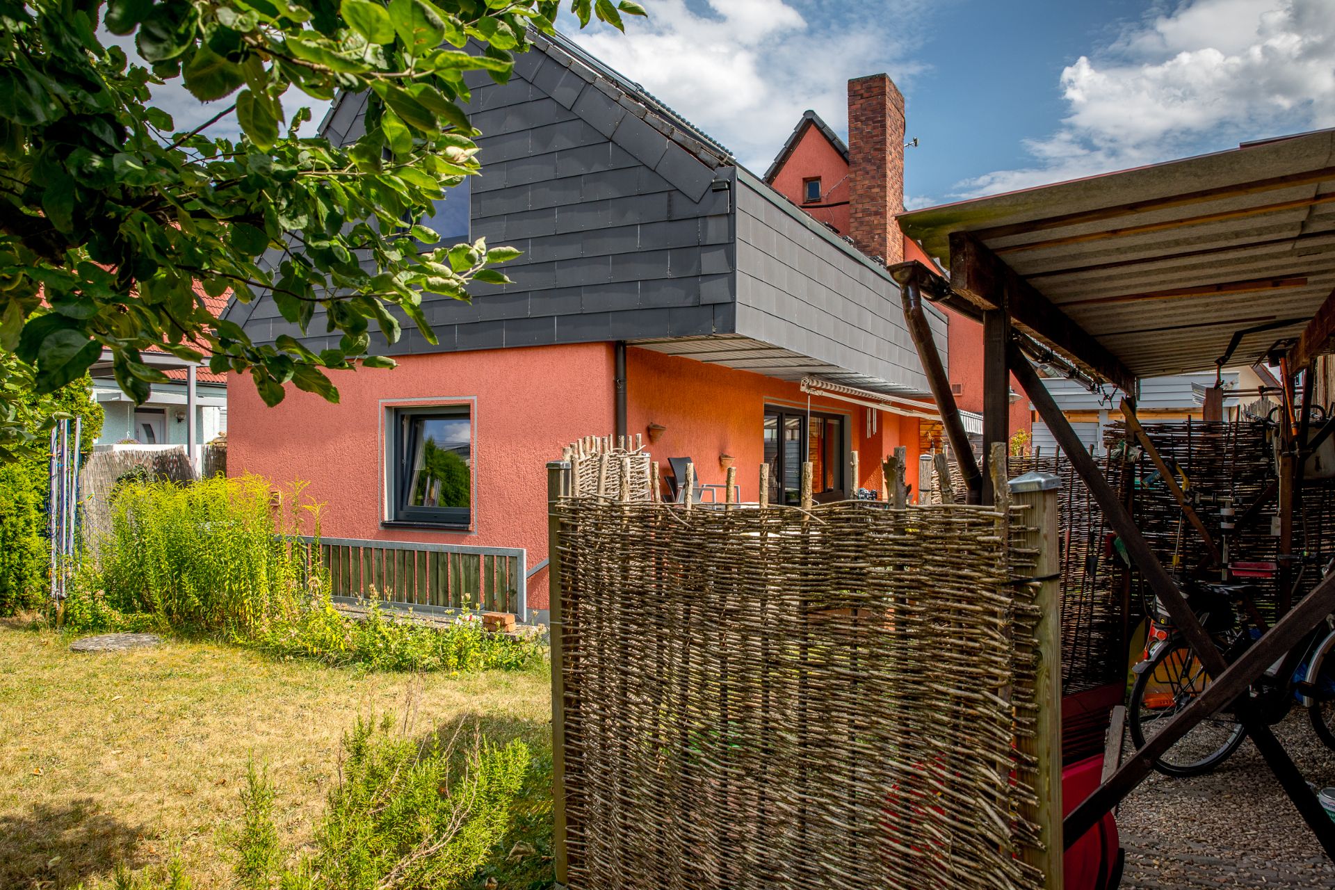 Haus mit roter Fassade, großem Fenster und Garten mit Zaun im Vordergrund.