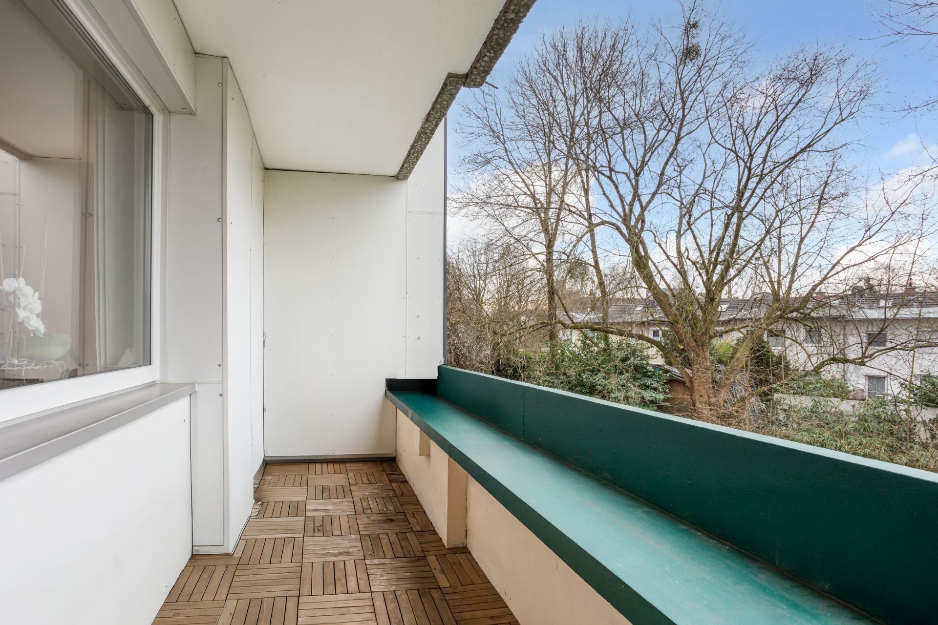 Überdachter Balkon mit Holzboden, Fenster links und Blick auf Bäume und Nachbarhäuser.