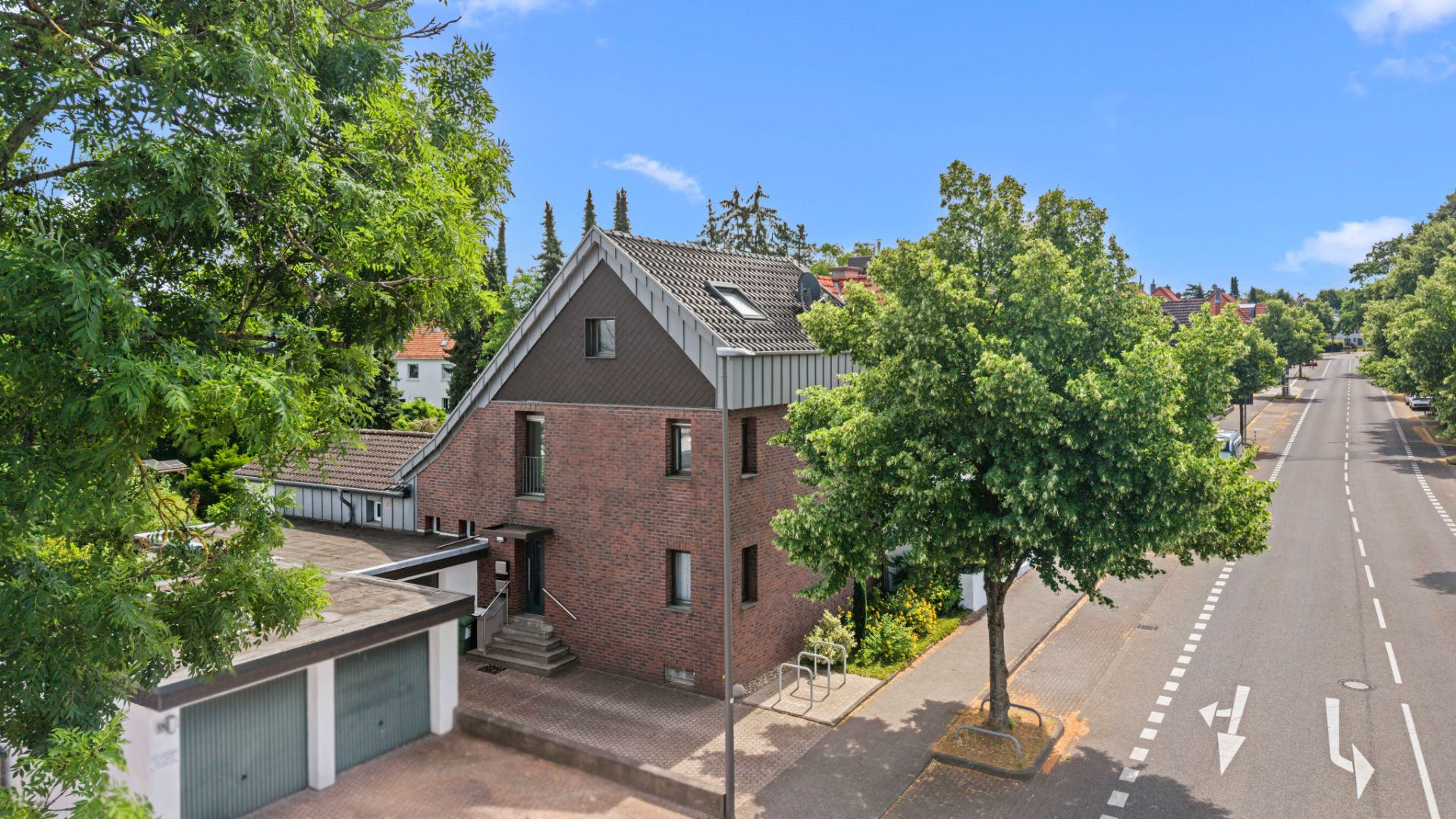 Zweistöckiges Backsteinhaus an einer Straße, umgeben von Bäumen und Garagen.