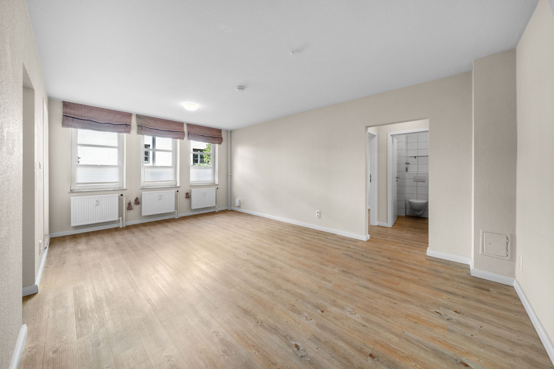 Helles, leeres Wohnzimmer mit drei Fenstern, Holzfußboden und Blick ins Badezimmer.