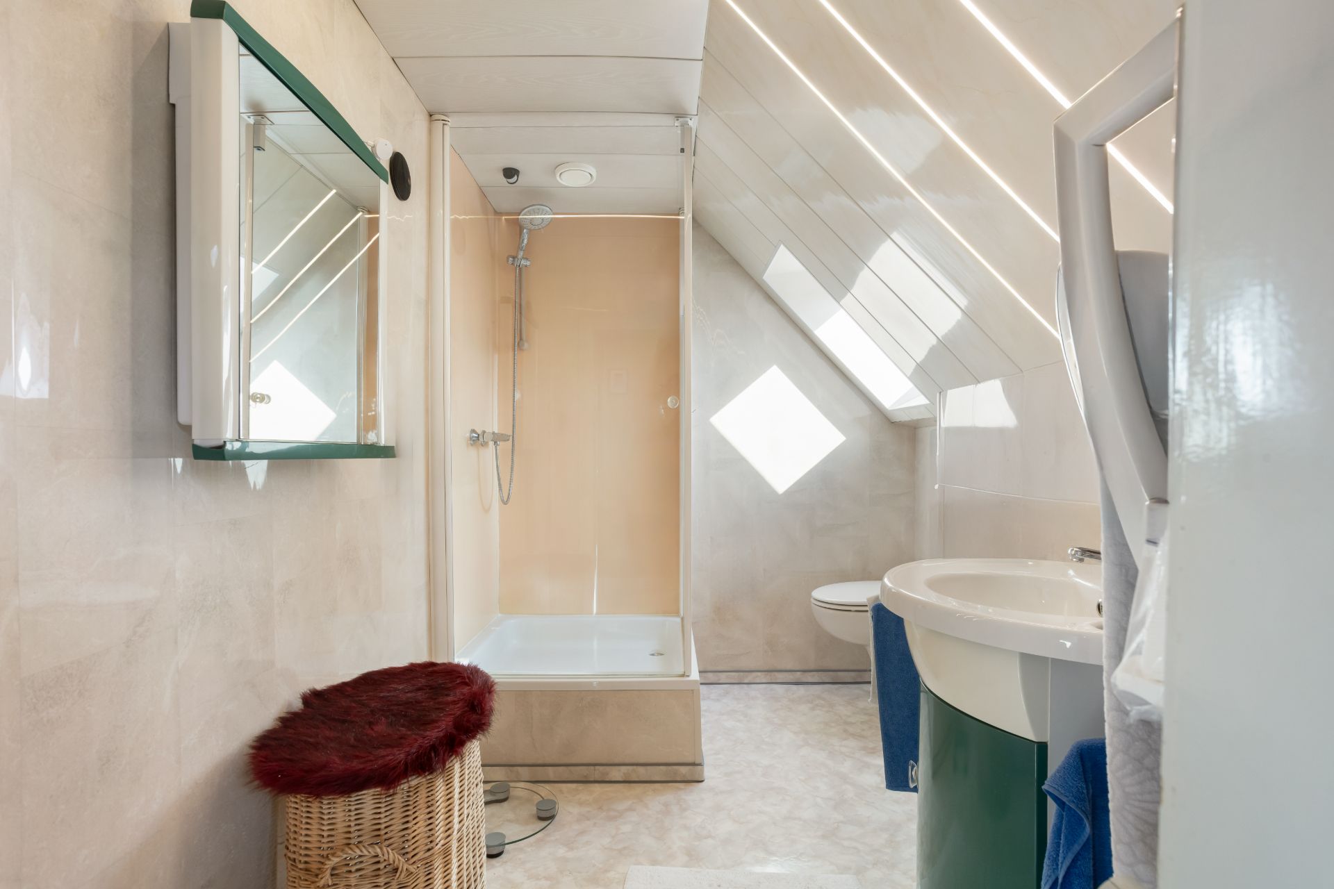 Modernes Badezimmer mit Dusche, Waschbecken, WC und Korb unter einer schrägen Decke mit Fenster.