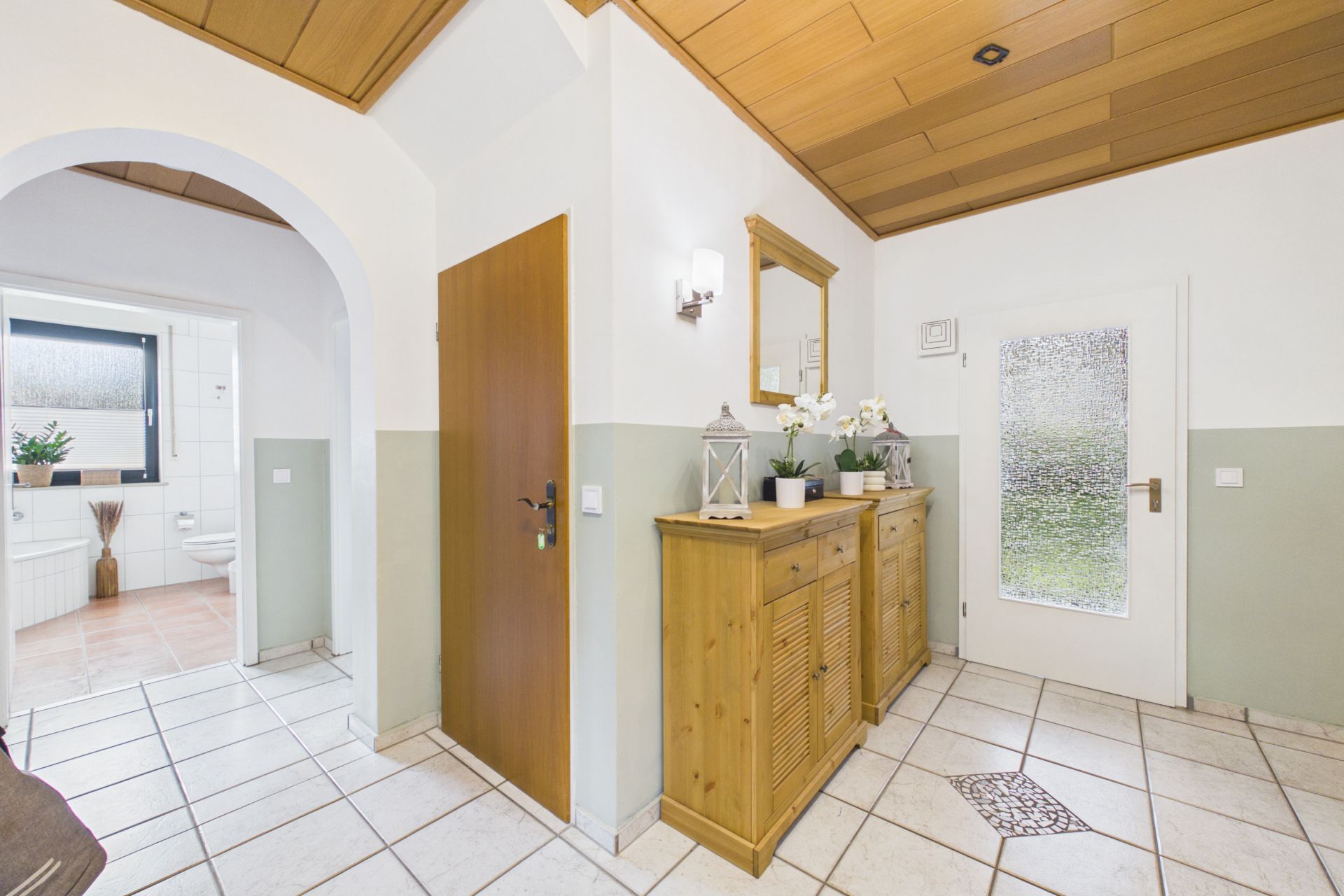 Heller Flur mit Fliesenboden, Holzdecke, Kommode, Spiegel und Blick ins Badezimmer.